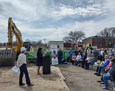 A speaker addresses a seated crowd with an excavator in the background.