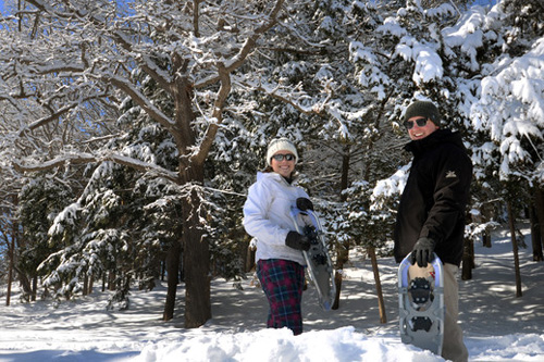 Two people smiling while holding snowshoes next to snow-covered pine trees.