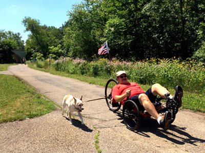 A dog on a leash with a man in a reclining bicycle in a park.