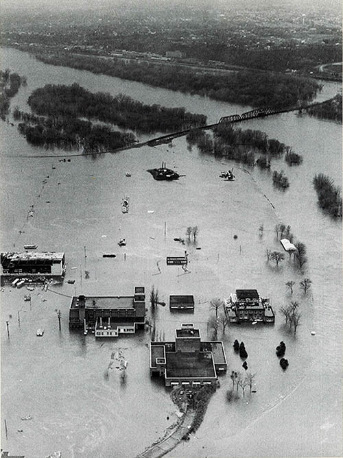 Black-and-white aerial image of a large area of land underwater next to a river.