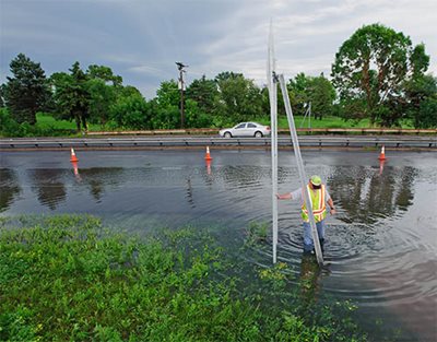 A worker in knee-deep water near a flooded road.