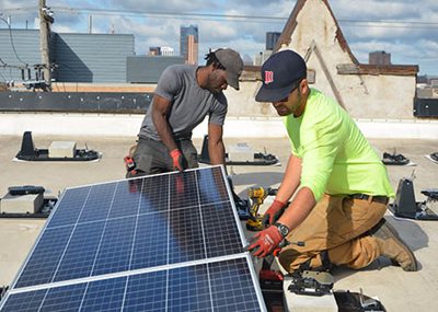 Two people with tools, kneeling next to a small solar panel.