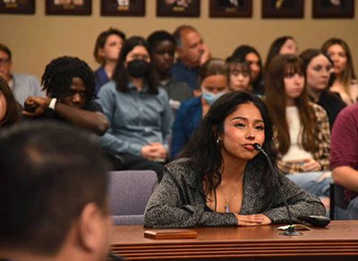 A young woman at a table, with crowd seated in back of her, speaks to the Met Council members.