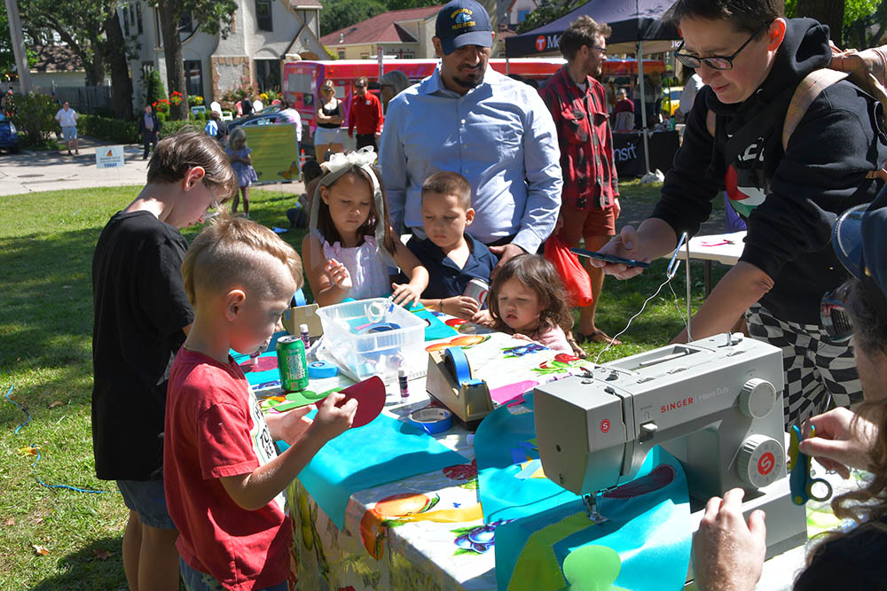 At table, children choose shapes to place on bright blue flags and place them with painter’s tape to the fabric. A man sews the flags on a sewing machine at the end of the table.