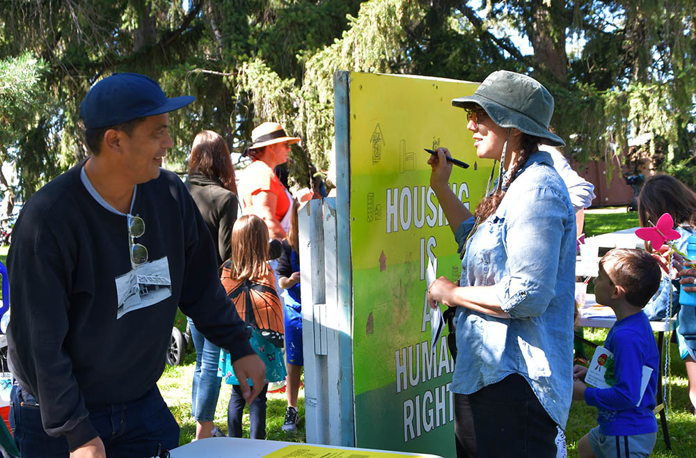 Man in baseball cap talks with woman using felt-tip pen to draw on multi-color poster that says, “Housing is a human right.”