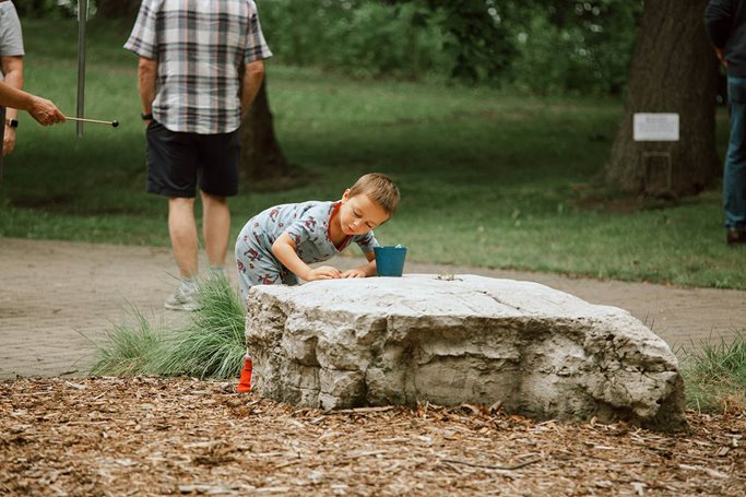 Boy leans over a vase sitting on a tree stump to hear the recorded sound coming out of it.