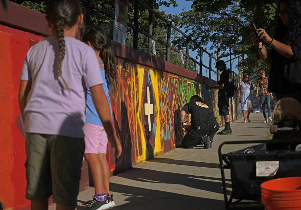 Two men and one child paint on a long mural, about four feet high, next to a sidewalk in the Raymond/University Avenue area of Saint Paul.