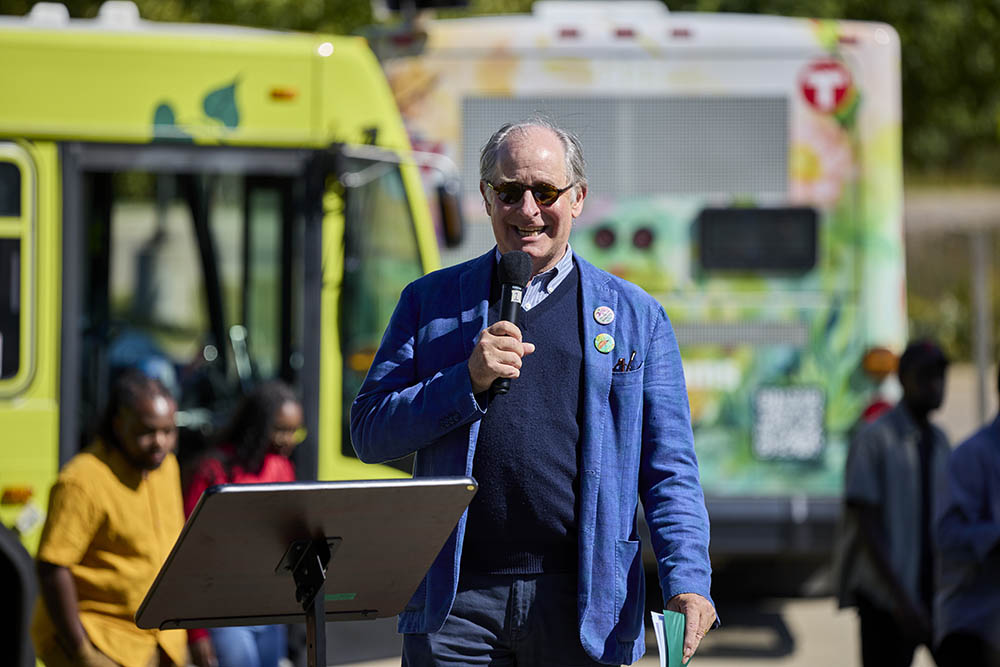 Charlie Zelle stands with microphone in front of two colorful Metro Transit buses.