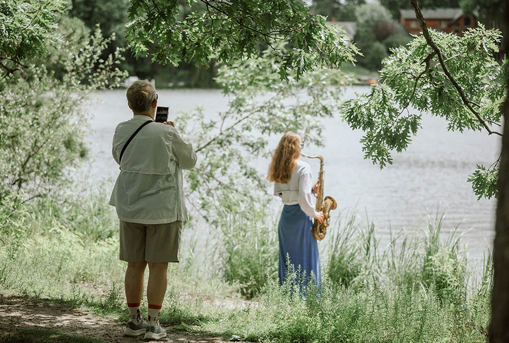 Woman plays saxophone at the shore of Silverwood Lake while another woman takes a photo of her.