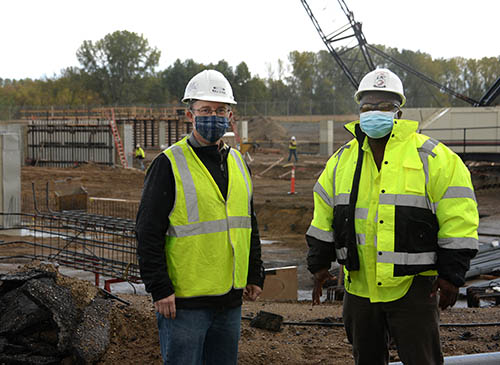 Two people in masks, hard hats, and reflective jackets at a construction site.