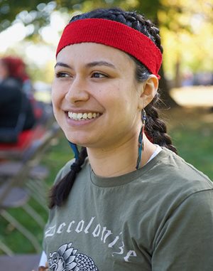 Close-up of smiling woman wearing feather earrings and a t-shirt that says “Decolonize.”