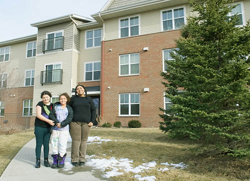 Three teens stand on a sidewalk in front of an apartment building.