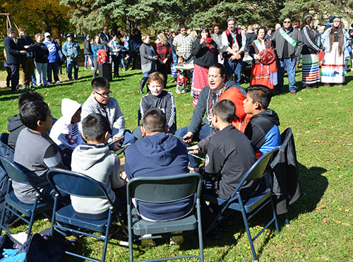 Eight Native youth and one elder sit in a circle around a large drum; other people listening are standing in the background.