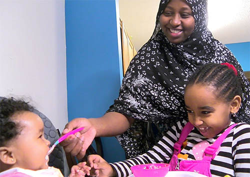 A woman feeding a child in a high chair, with another child nearby.