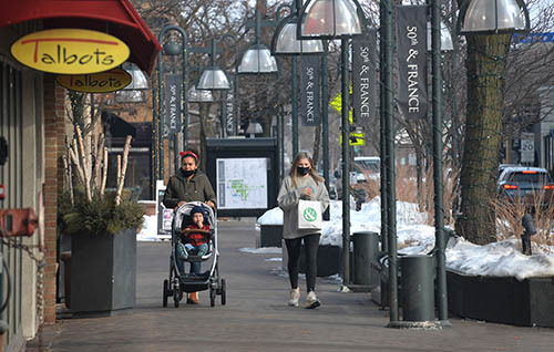 Two people, one pushing a stroller, on a sidewalk near stores.