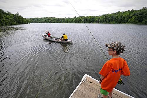 Two people in a canoe and a child fishing off a dock.