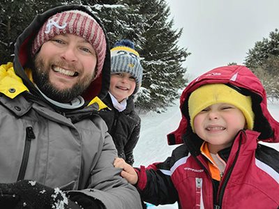Selfie of Anthony and two children in front of snow-covered pine trees.
