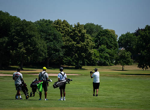 3 people watching a 4th person swing on a golf course on a sunny day.