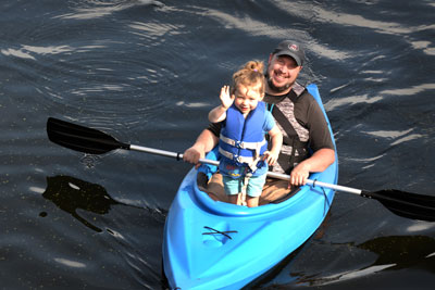 An adult and child at rest in a kayak. The child is waving at the camera.
