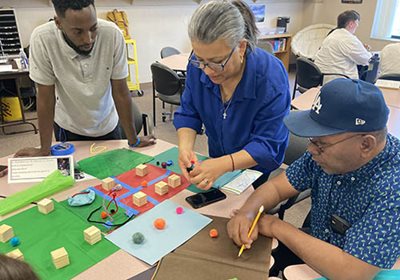 Three people use craft materials to show a neighborhood layout at a table during a community meeting.