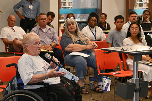 Woman in wheelchair speaks into microphone as large group of people seated nearby listen.