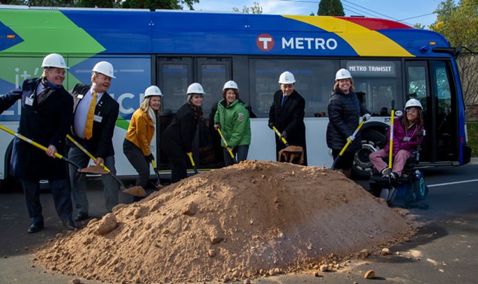 7 people standing and 1 person in a wheelchair, all wearing hard hats and holding yellow shovels to scoop dirt for the groundbreaking ceremony.