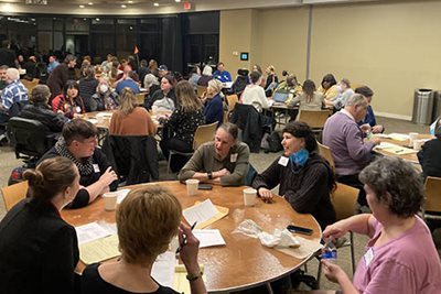 A large room with several round tables, with people filling every seat at the table.