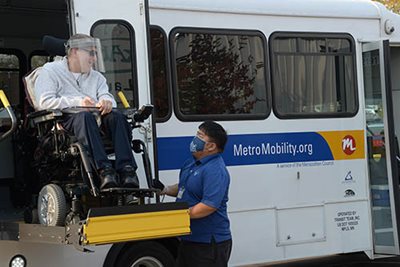 A Metro Mobility customer wearing a mask in a wheelchair, assisted on the lift by a Metro Mobility driver in a mask.