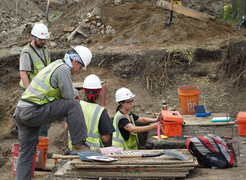 Four people in green vests and helmets in a dig site.
