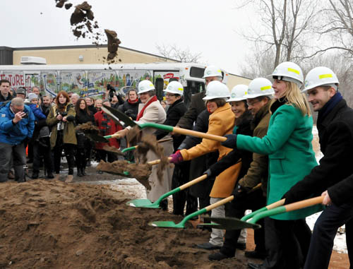 Eight people wearing hard hats, with green shovels. One person is tossing sand into the air.