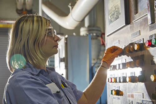 A woman in safety glasses stands in front of a control panel with many buttons.