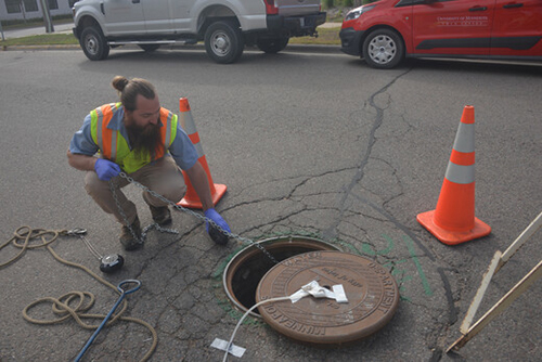 Worker in a fluorescent vest at street level checks instruments in a maintenance hole in the street.