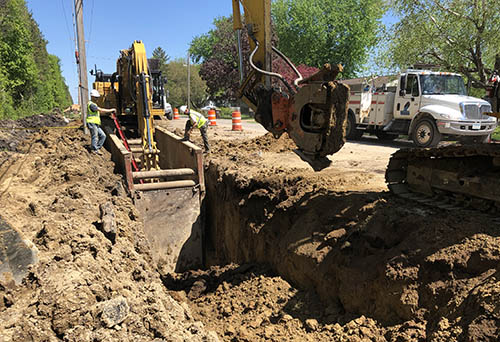 People working with equipment on a trench next to a road.