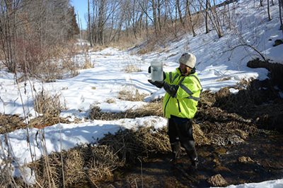 A worker in a safety vest holding up a container with water in a waterway in winter.