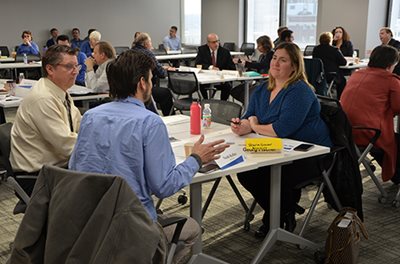 Three people talking at a table, with other small groups at tables in the background.