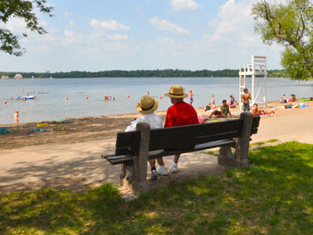 Two people sitting on a bench wearing hats, looking onto a clean lake and beach with several people.
