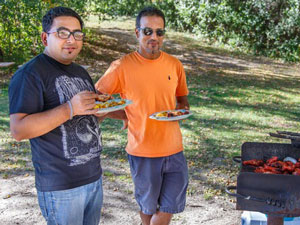 Two people hold plates of food next to a grill in a picnic area at a regional park.