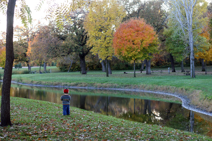 A child in a sweater and a knitted pumpkin hat next to a creek among trees with bright colors.