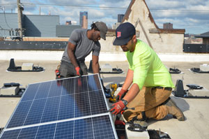 Two people install a solar panel on a roof using powered tools. It’s a partly cloudy day and both people are wearing a hat and work gloves.
