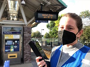 A worker in a mask with a digital device waiting on a train platform to talk to passengers.