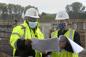 Two workers in safety vests, hard hats, and masks look at construction plans.