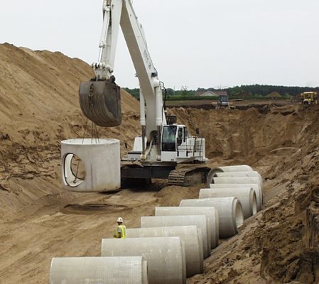 Image of previous sewer construction project in process, showing an excavator lifting a large section of pipe with a hole through it.