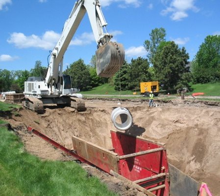 Image of previous sewer construction project in process, showing an excavator on the edge of a hole with a construction structure at the bottom. The excavator is lowering a piece of pipe into the hole.