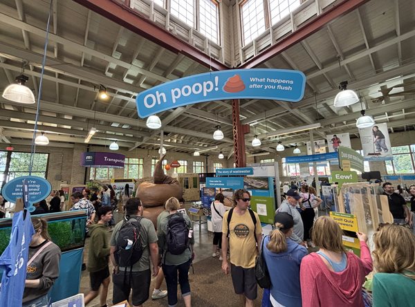 Visitors move through the Oh poop! exhibit, stopping to take a sniff test (right). Overview of the Oh poop! exhibit with a hanging sign and many people.