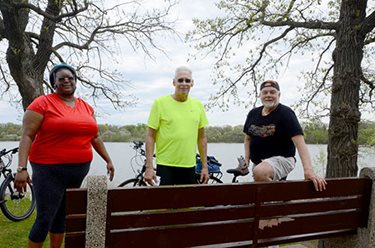 Three people stand near a bench with their bikes, lake in the background.