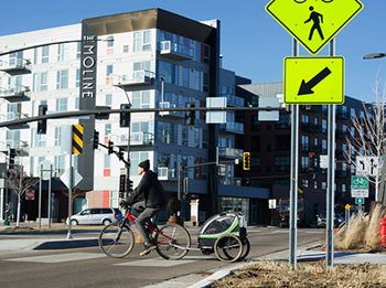 Man on bicycle with trailer is crossing street near the Hopkins Station; newer apartment building The Moline is in the background.
