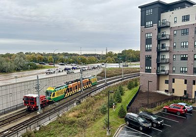 Special locomotive car pulls two light cars on the tracks near a highway and a tall apartment building.