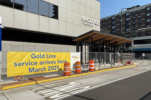 Fence surrounds sheltered benches and station platform. Banner on fence reads Gold Line service arrives here March 2025.